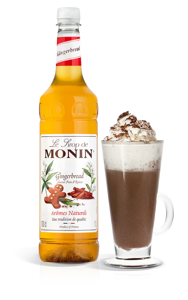 Monin gingerbread syrup bottle with a glass of hot chocolate topped with whipped cream and nuts on a white background