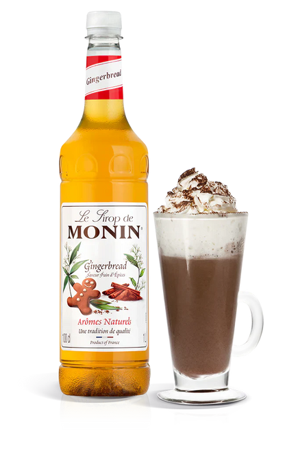 Monin gingerbread syrup bottle with a glass of hot chocolate topped with whipped cream and nuts on a white background